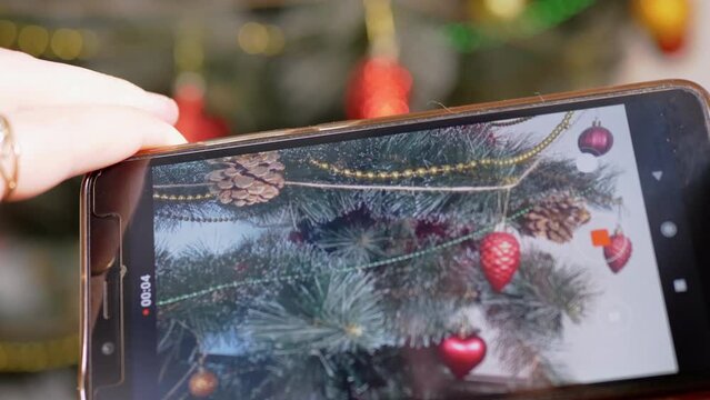 Female Hands are Shooting a Video of Christmas Tree on a Smartphone. Blogger takes pictures, shoots Christmas tree decorations on a mobile phone camera. Preparing New Year. Photographing event. 4K.