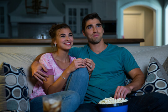 Young couple sitting on couch watching a movie at home with a bowl of popcorn 