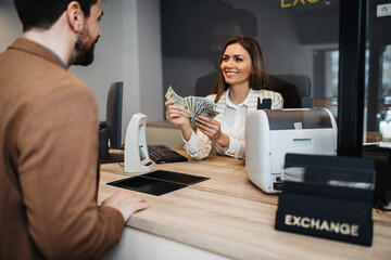 Attractive European businesswoman working in exchange office. She is giving cash money to male customer. - obrazy, fototapety, plakaty