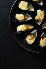 baked mussels in shells with creamy sauce poured with oil on a black plate on a dark background