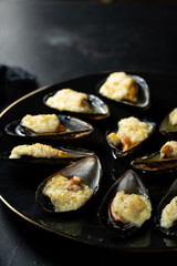 baked mussels in shells with creamy sauce poured with oil on a black plate on a dark background