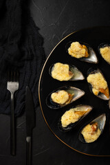 baked mussels in shells with creamy sauce poured with oil on a black plate on a dark background