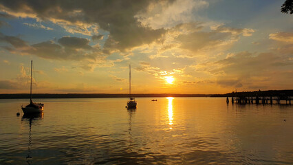 Fototapeta na wymiar Romantische und Entspannende Landschaft. Boots in Sonnenuntergang am Ammersee. Glühende Sonn spiegelt sich im See, Himmel voller Wolken und Farben. Feurig orange leuchtende Sonne am Horizont. 