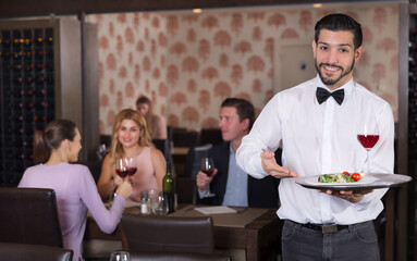 Polite waiter holding tray at restaurant with customers his behind