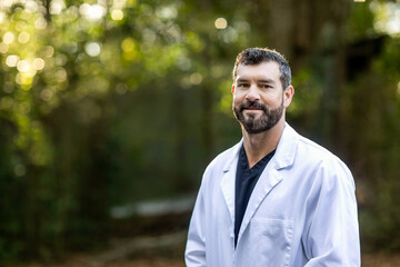 A doctor with dark hair and a beard in a white lab coat standing outside in a natural green...