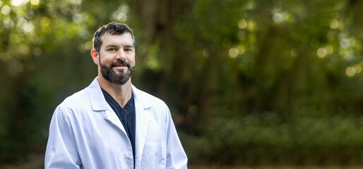 A doctor with dark hair and a beard in black scrubs and a white lab coat standing outside in a...