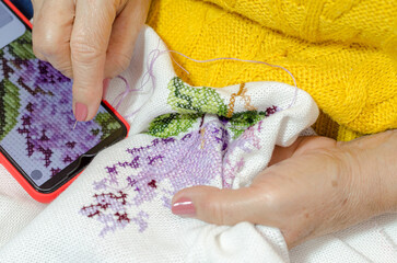 Older adult woman embroidering with a cross-stitch needle. Close-up. Technologies of different...