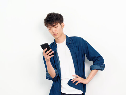 Portrait Of Handsome Chinese Young Man With Curly Black Hair In Blue Shirt Posing Against White Wall Background. Looking At His Smartphone In Hand With Another Hand On Waist, Studio Shot Front View.