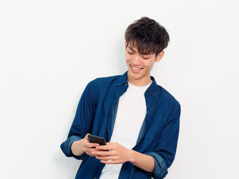 Portrait of handsome Chinese young man with curly black hair in blue shirt posing against white wall background. Smiling and looking at his smartphone holding in both hands, front view studio shot.