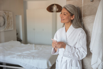 Smiling mature lady in bathrobe with smartphone in hotel room