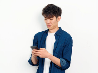 Portrait of handsome Chinese young man with curly black hair in blue shirt posing against white wall background. Looking at smartphone in both hands with disgusted expression, front view studio shot.