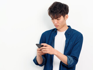 Portrait of handsome Chinese young man with curly black hair in blue shirt posing against white wall background. Looking at smartphone in both hands with serious expression, front view studio shot.