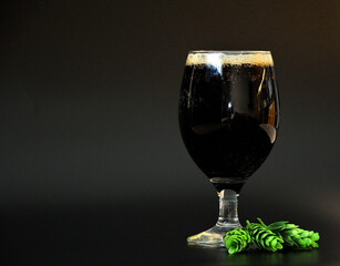 A glass of dark fresh beer with white dense foam and green fruits of ripe hops on a black background.