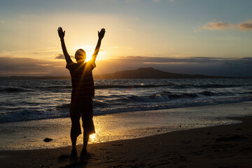 Fototapeta na wymiar Silhouette of a man with open arms greeting the sun rising over Rangitoto Island, Milford Beach, Auckland.