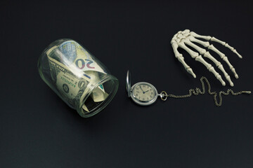 Banknotes of different money in a glass jar, a round pocket watch and a skeleton palm on a black background