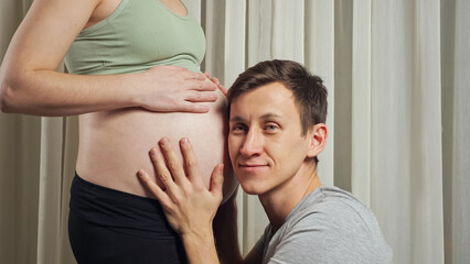 Brunet young husband kisses and listens to unborn baby moves in stomach of pregnant wife wearing...
