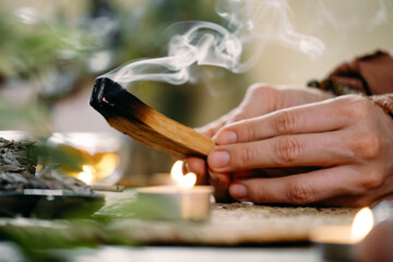 Woman hands burning Palo Santo, before ritual on the table with candles and green plants. Smoke of smudging treats pain and stress, clears negative energy, meditation wooden stick