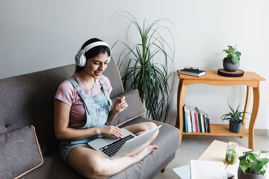 young latin Woman student with headphones using computer in a video call or online class while sitting on sofa at home in Mexico	