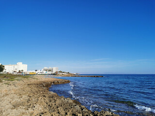 A stone shore made of solidified lava, in the distance a small sparsely populated sandy beach with sun loungers and sun umbrellas in the bay of the Mediterranean Sea on a sunny day.
