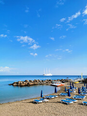 Obraz premium Sandy beach in a bay with sun loungers and umbrellas, in the Mediterranean Sea the largest sailing yacht in the world, an eight-deck motorsailer against a blue sky with clouds.