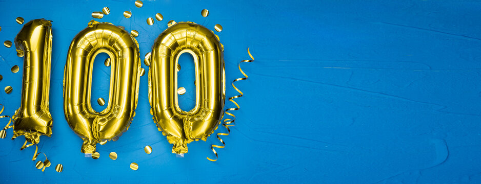 golden yellow foil balloon on blue concrete background number one hundred. Birthday or anniversary card with 100 inscription. Anniversary celebration.