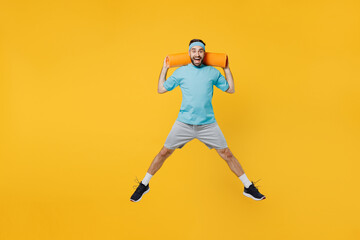 Naklejka na ściany i meble Full body overjoyed excited happy young fitness trainer instructor sporty man sportsman wear headband blue t-shirt hold yoga mat jump high isolated on plain yellow background. Workout sport concept