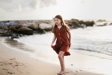 Cute long-haired European baby girl in a dress on the sea coast. Girl and stones at sunset on the ocean.