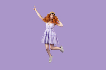 Full-length photo of a joyful funny redhead girl in summer short dress with a straw hat jumping and pointing up with a finger.