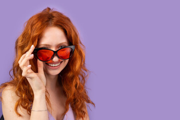 Young redhead woman in spring summer dress smling widely taking off her red sunglasses on purple background and copy space for advertising.