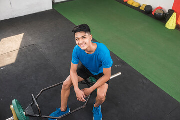 A satisfied young man smiling while sitting on a hex trap bar after training at the gym. High on endorphins. A successful and fulfilling workout session.