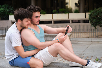 Two young men using the smartphone sitting on a bench in the street. Gay couple.