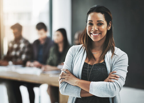 Shes confident about her teams abilities. Portrait of a cheerful businesswoman posing with her arms folded in a modern office with her colleagues in the background.