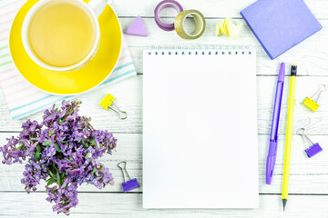 Fototapeta na wymiar Notebook (sketchbook) with a blank sheet of paper for writing, pen, pencil, yellow mug with tea on a saucer, stationery and lilac spring flowers on a white wooden background. Space for text. 