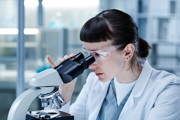 Young female doctor in protective gloves looking through the microscope to examine the new virus in laboratory