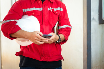 An engineer or manager in red uniform is holding smartphone and white safety helmet during take group meeting with team staff. Industrial working people in action photo. Selective focus at hand part.