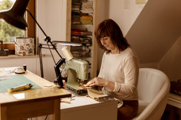 Caucasian brunette woman sewing fabric with a vintage, retro sewing machine. Fashion, creation and tailoring. Process of sewing in atelier or workshop. Special hobby. Cozy interior and warm colors.