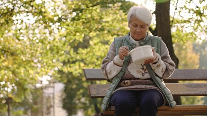 Fototapeta premium Old white-haired homeless woman eating warm meal thanks to the charity volunteers that help people in need. High quality photo