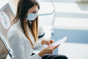 Female wearing protective mask is standing at the window in the airport terminal awaiting the...