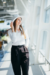 Fototapeta premium Cool young woman in boater and white dress smiles and looks into camera outside. Tourist holds passport and poses on crosswalk near airport.
