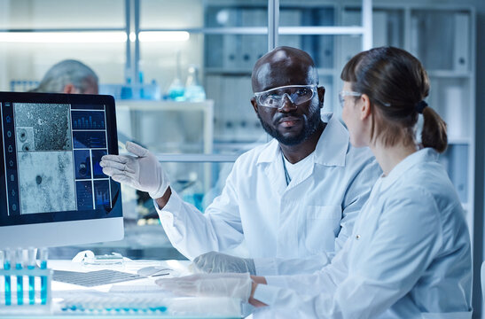 Diverse Couple Of Scientists Discussing Image Of Bacterium On Computer Monitor Together Sitting At Table