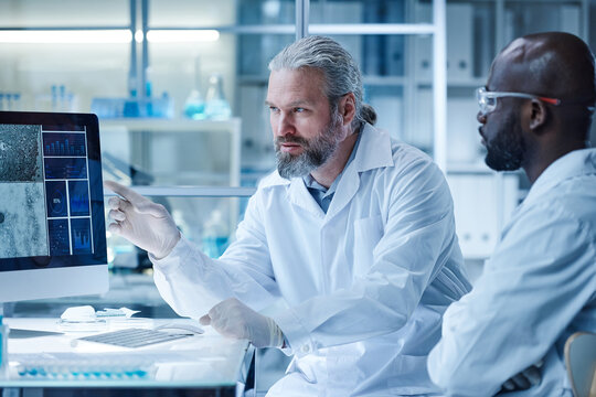 Bearded male doctor pointing at computer monitor with images of virus and talking to his colleague during teamwork in laboratory