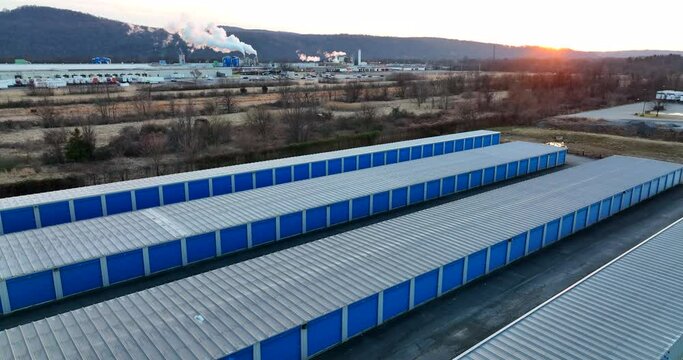 Self storage units with blue garage doors. Rising aerial at sunrise, sunset. Factory in distance. American rental units.