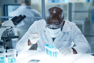 African scientist in mask taking analysis with pipette from flask during medical research in...