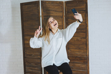 Emotional young woman in a white cotton shirt gesticulates and looks at the screen of a modern...