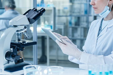 Close-up of woman in protective gloves and lab coat using digital tablet to examine the samples on the screen after researching them on microscope