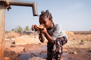 Young black girl greedily drinking fresh water from a pipe somewhere in an African dryland; global...