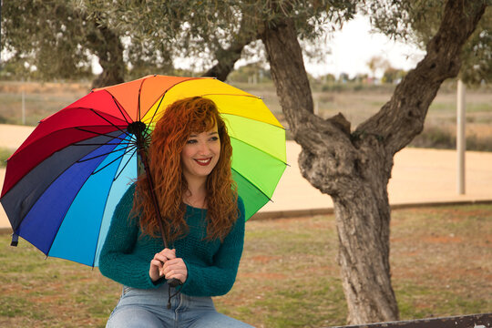 Portrait of young woman, red hair, freckles, with a rainbow umbrella, sitting on a wooden bench, happy, in an outdoor park. Concept color, happiness, well-being, fun, rain.