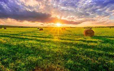 Gordijnen Scenic view at picturesque burning sunset in a green shiny field with hay stacks, bright cloudy sky , trees and golden sun rays, summer valley landscape © Yaroslav