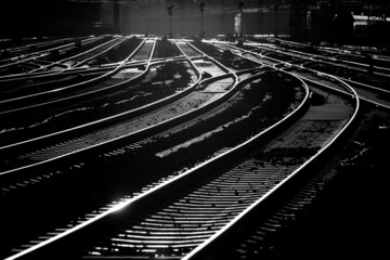 Bent parallel railway tracks at Dortmund Main station Germany with switches, signals, thresholds, catenary at sunset. Black and white greyscale with high contrast. Technical infrastructure for trains.