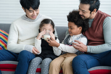 Happy young Asian gay couple with diverse adopted children, African and Caucasian, drinking milk and sitting on sofa at home. Older boy with milk mustache cheer up little girl drink milk. Lgbt family